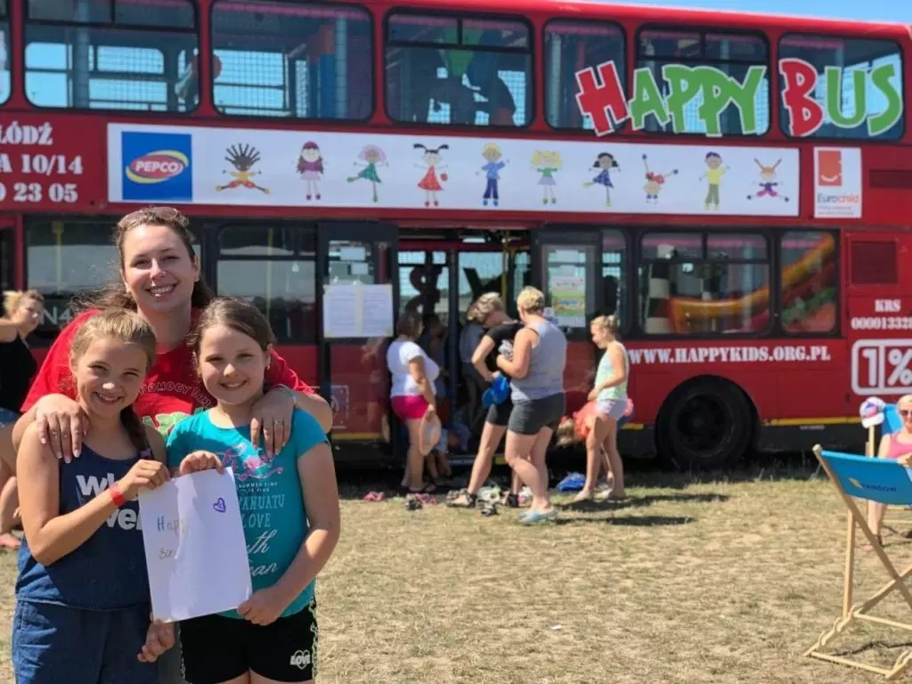 Naine ja kaks last poseerivad punase kahekorruselise HAPPY BUS bussi ees, laps hoiab joonistust.